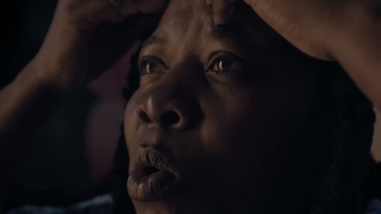 Foto del rostro de una mujer de tez negra que lleva las manos en la cabeza y los labios entreabiertos como si pronunciara alguna palabra de asombro.